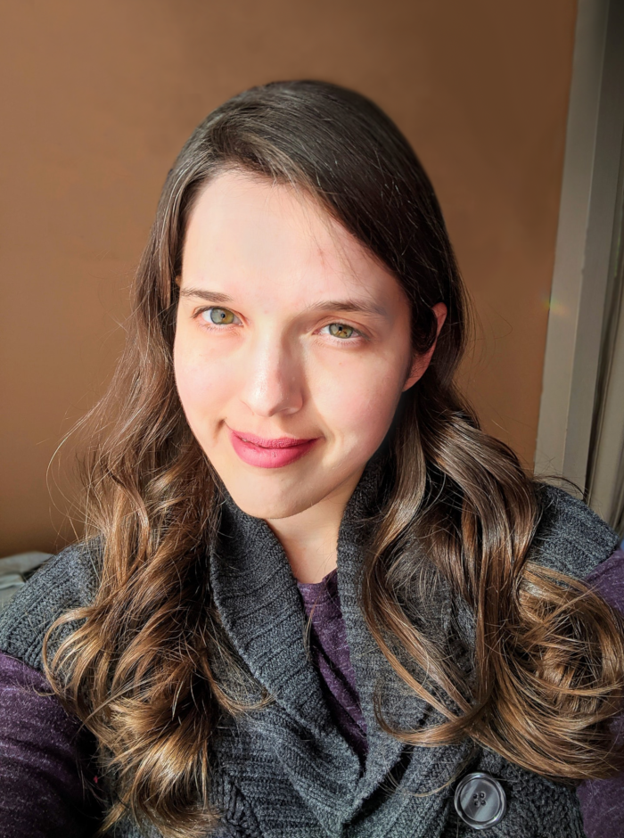 Headshot of Aili. She's wearing a purple shirt and a gray sweater vest. She is a white woman with hazel eyes and brown hair, which is looking uncommonly nice on this day. She's smiling slightly